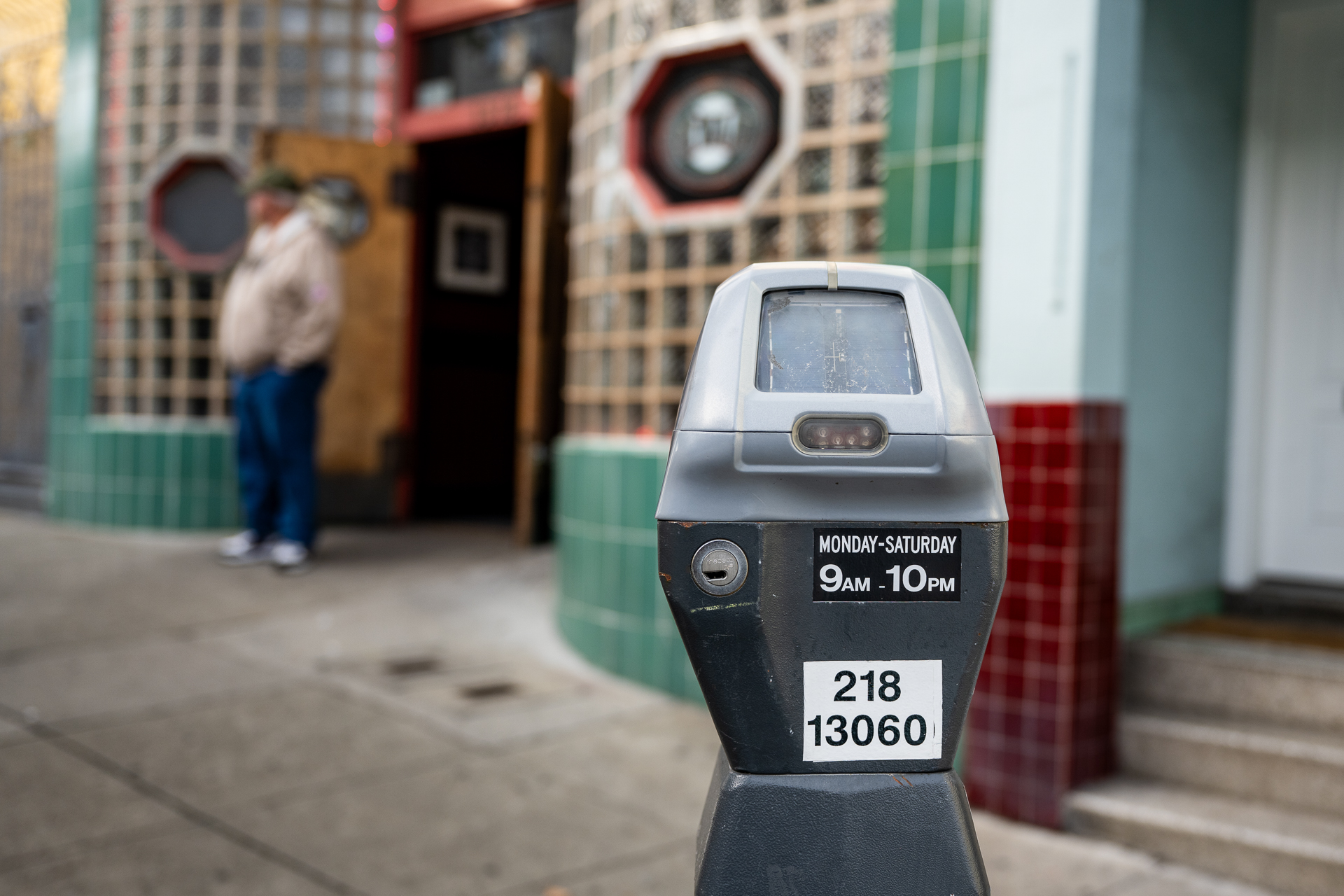 A diamond shaped metal parking meter with a digital face on a city street with a sticker that says, 'Monday - Saturday 9am-10pm.'