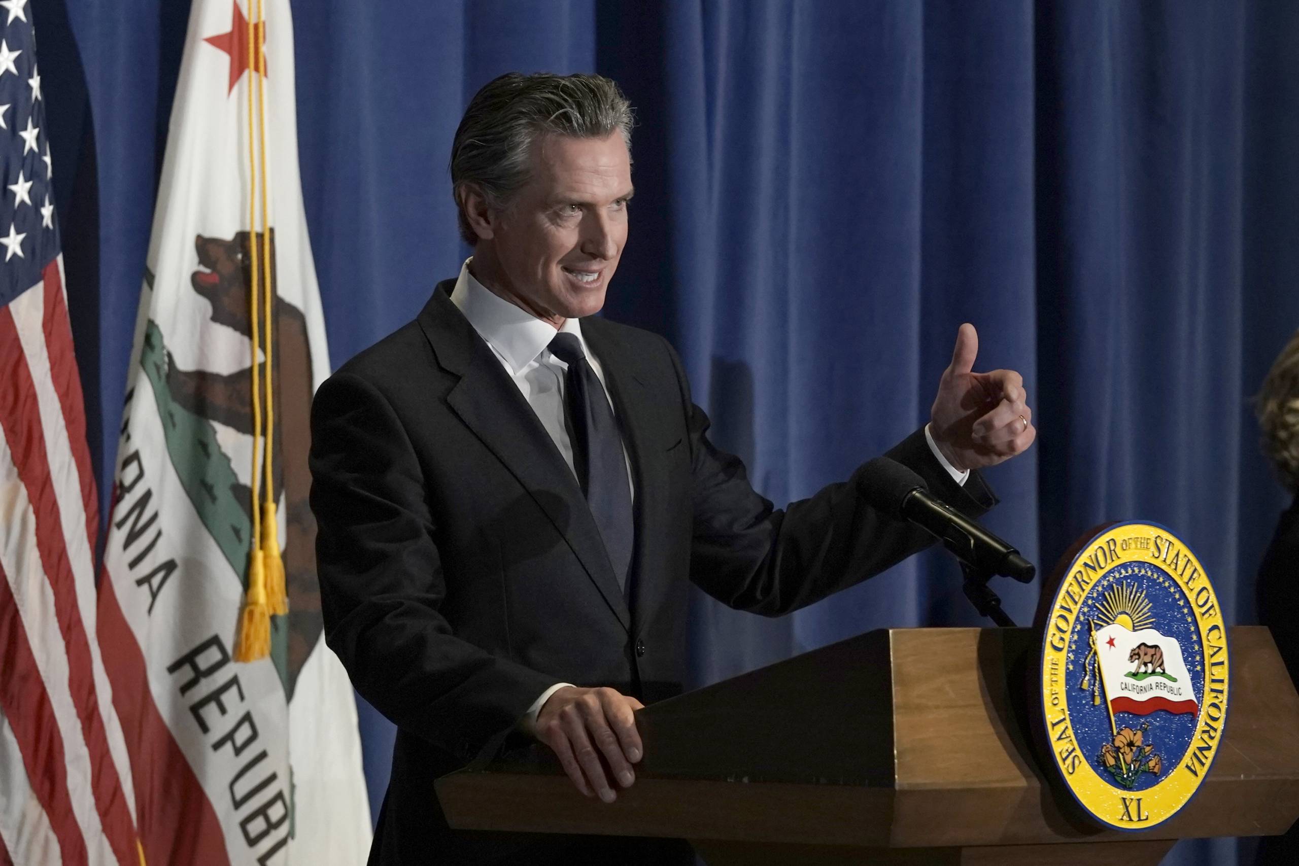 A white man in a business suit stands at a podium with his left hand motioning.