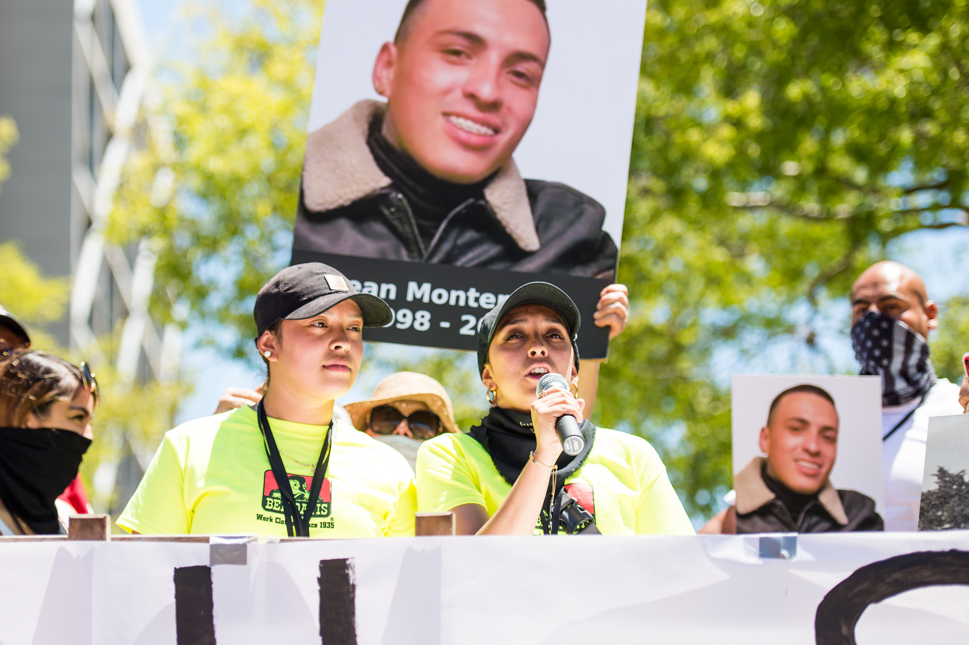 A rally is taking place with two women at a podium. One holds a microphone. A large photo of a man is behind them.