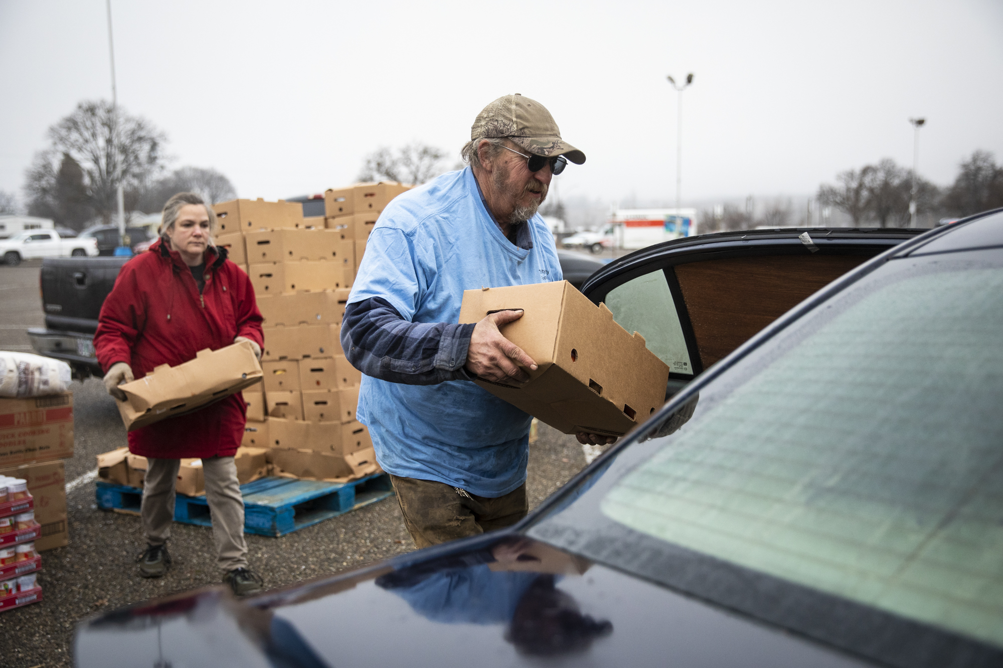 A man wearing a blue shirt, green hat and sunglasses holds a cardboard box of food to be moved into a car, with another man and several boxes behind him.