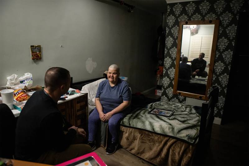 A woman with little hair and a blue outfit sits on a bed talking to a man.