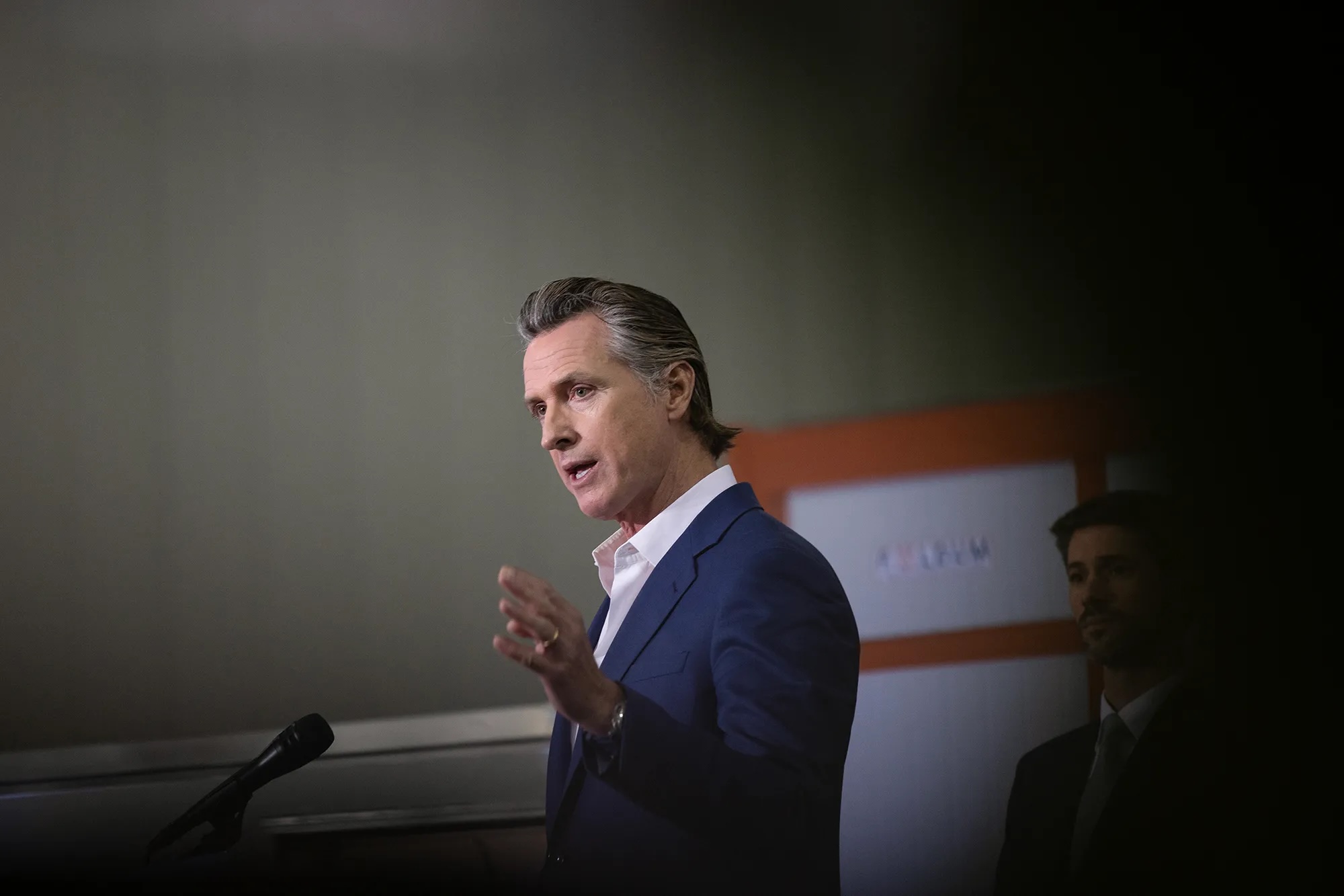 A white man in a blue suit and tieless white shirt gestures as he speaks.