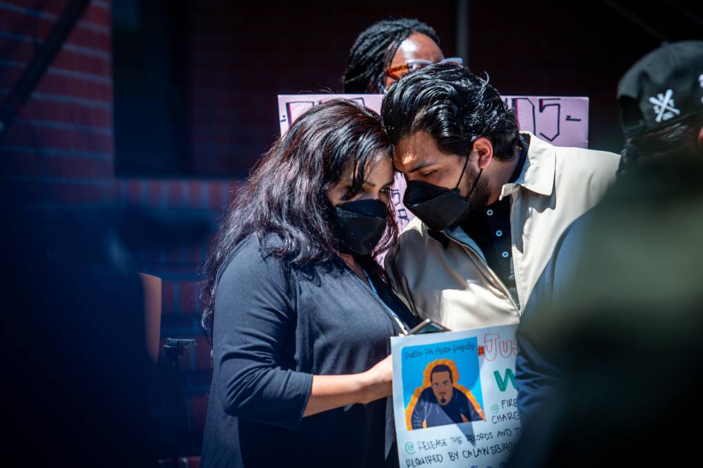 A Latina woman and a Latino man touch heads as they mourn with masks on.