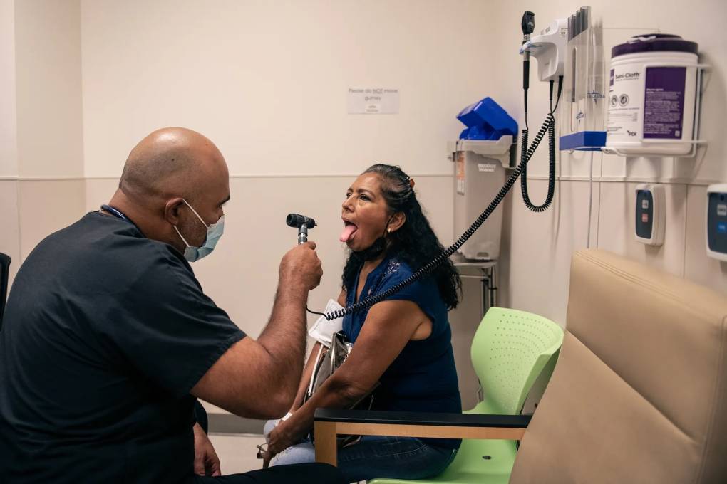 A male doctor examines a seated female patient who is sticking out her tongue in an exam room.