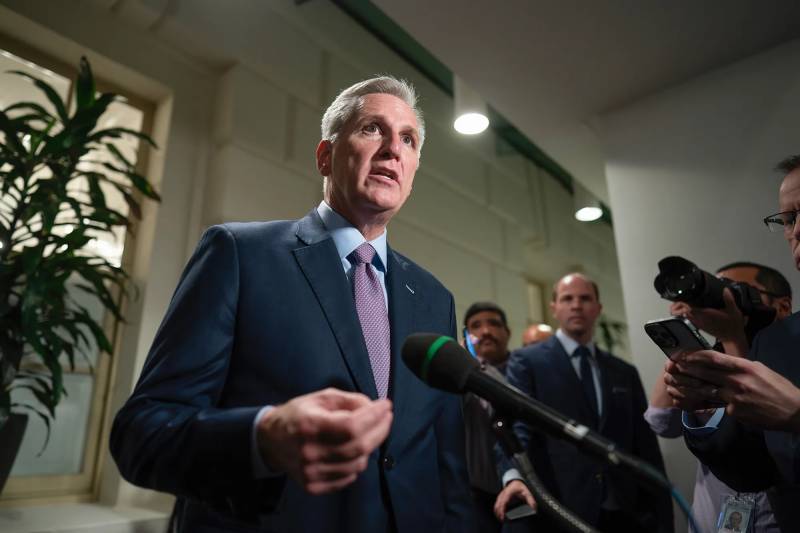 Kevin McCarthy speaks into a microphone at a press event surrounded by people.
