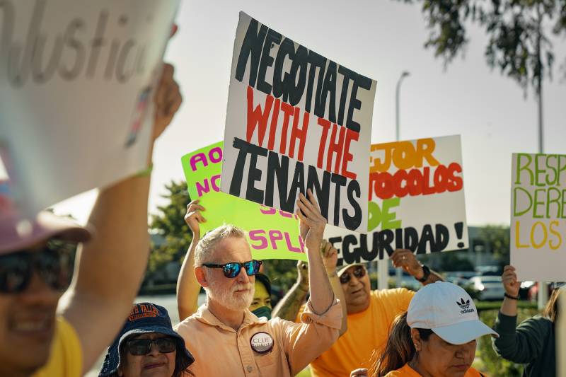 A man wearing an orange shirt and sunglasses holds up a sign in a crowd that reads "Negotiate with the Tenants."