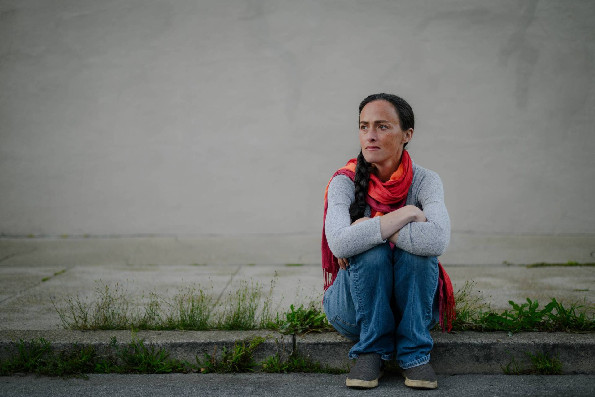 A woman wearing a scarf sits on a curb with her arms crossed.