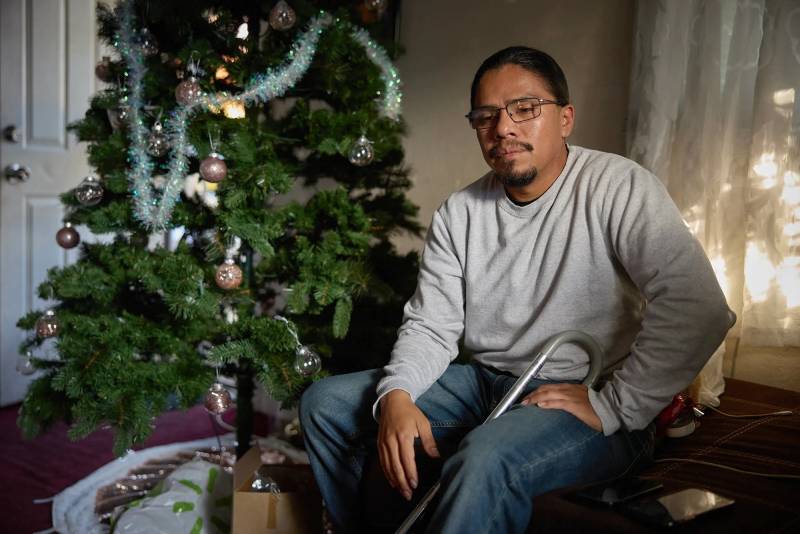 A Latino man with glasses sits next to a Christmas tree in a living room.