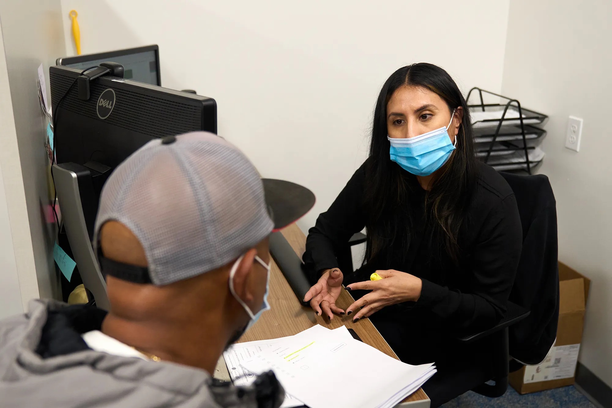 A young woman wearing a face mask sits at her desk and talks to a man in a baseball cap. A small stack of paper is between them.