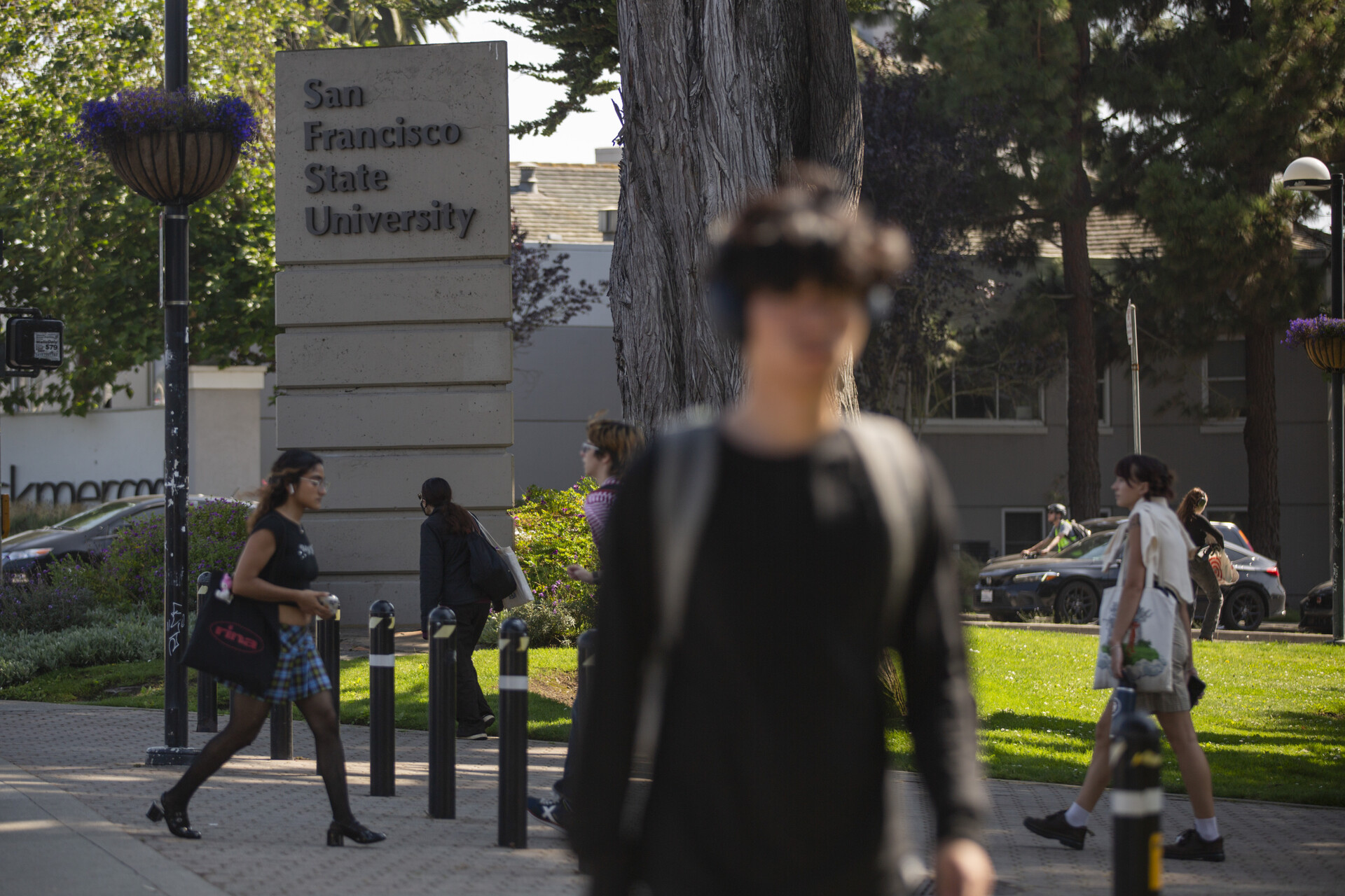 Many students, wearing backpacks, walk across a college campus. A large sign says, 'San Francisco State University.'