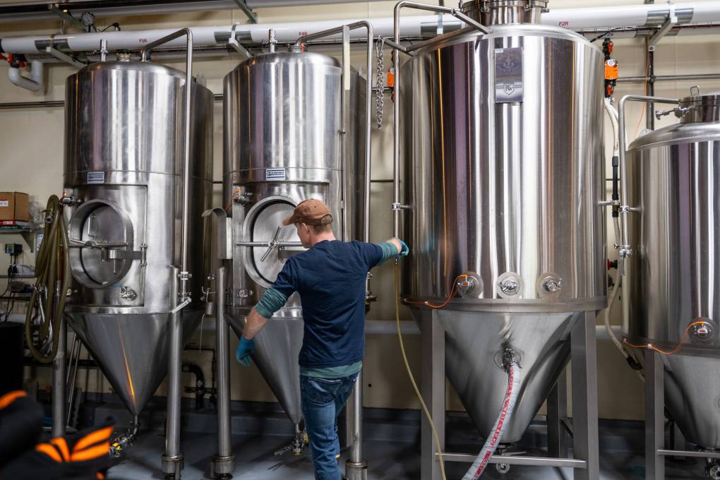 A person stands in front of three stainless steel fermentation tanks.