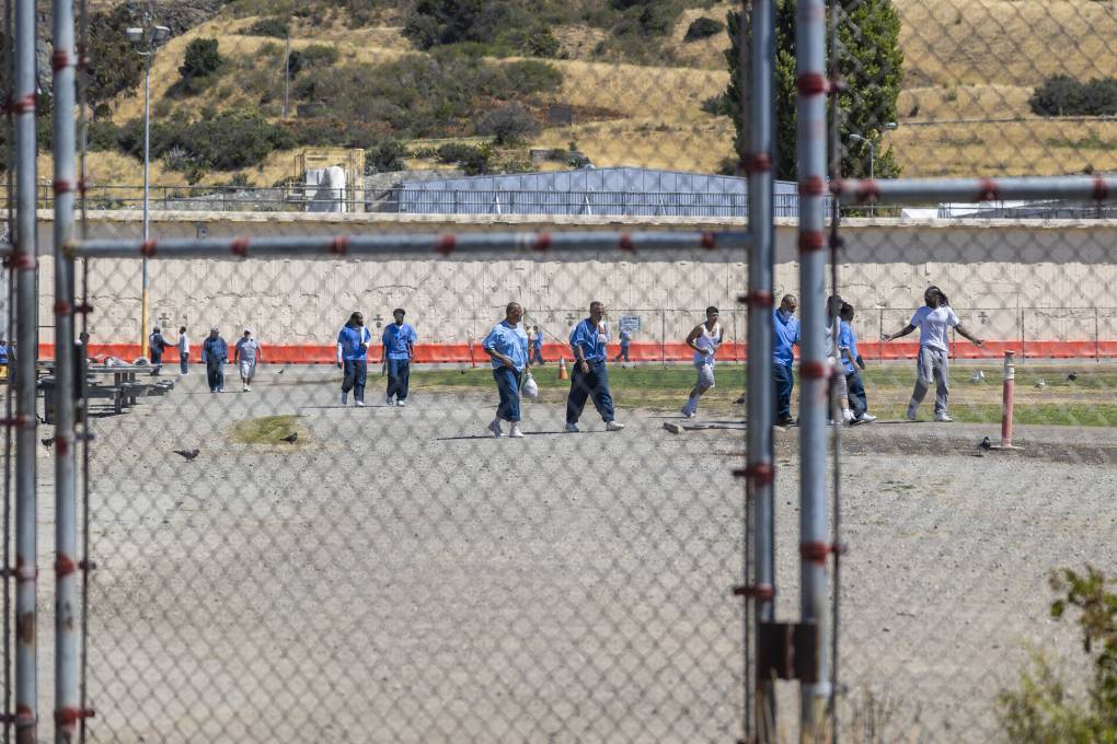Men in blue uniforms walk around a large asphalt yard behind a locked fence.