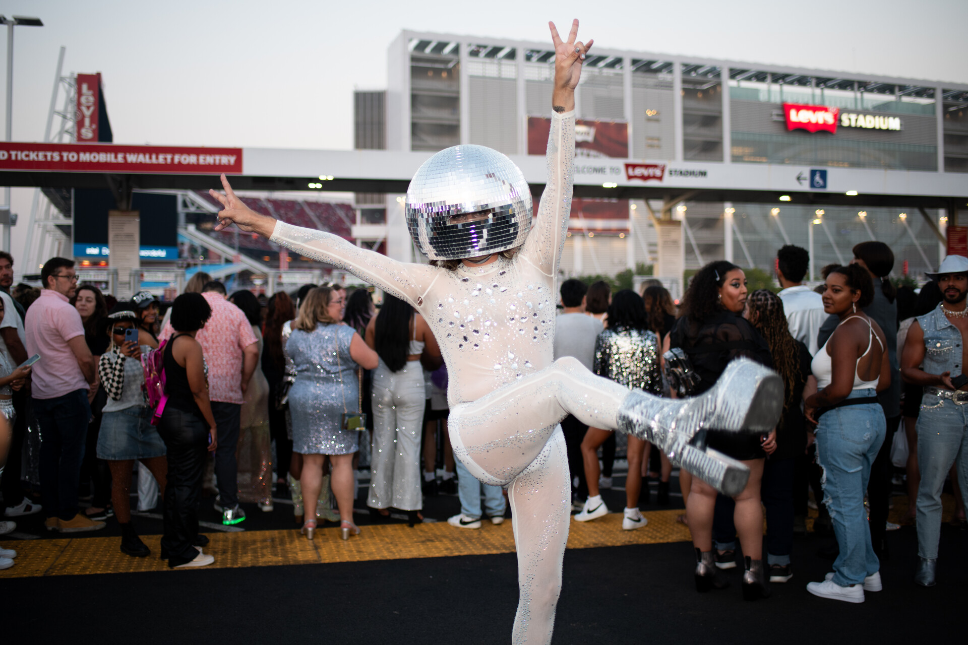 A person wears a disco ball on their head with a white sequined body suit.