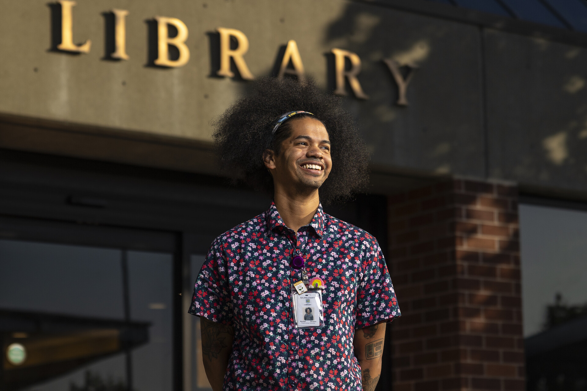 A person with their hair in an afro stands in front of a building with the word "Library" written over the entrance.