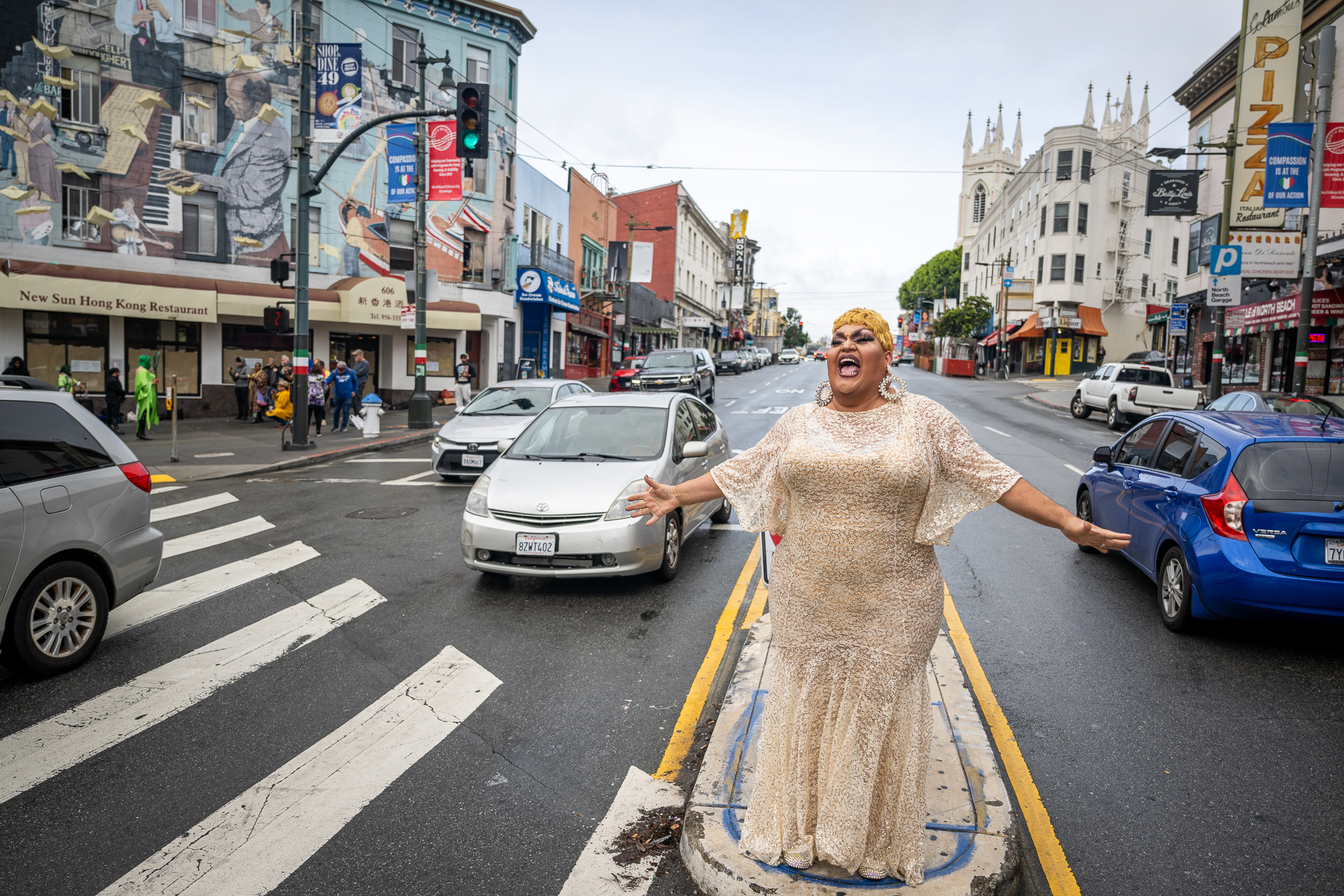 A drag performer in a white dress singing on a traffic island as cars go by.
