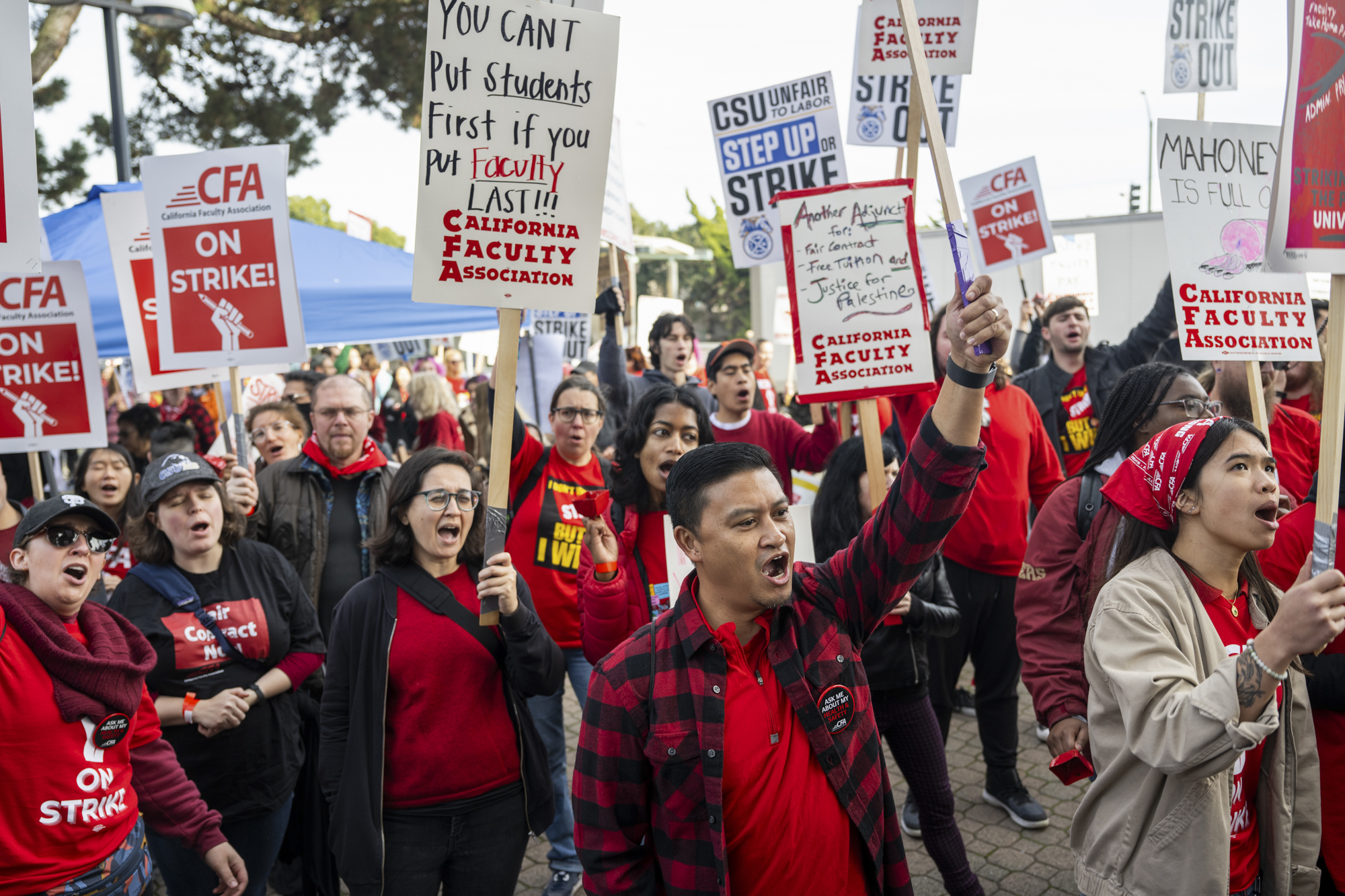 A group of people in red shirts cheer and carry picket signs in an outdoor setting.