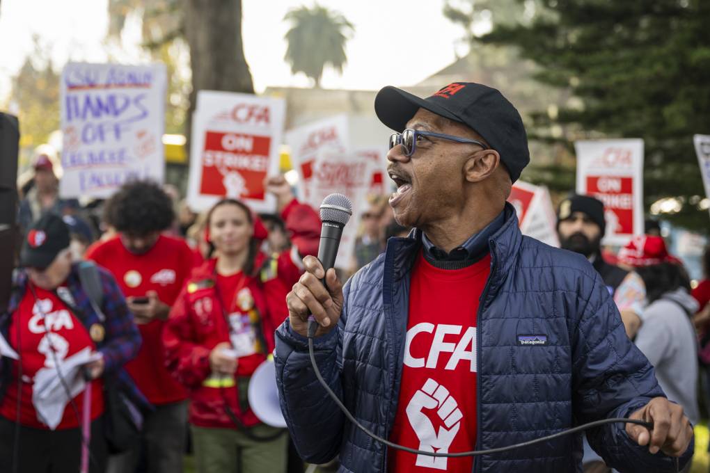A person in a baseball cap speaks into a microphone in front of a group of people in red shirts carry picket signs in an outdoor setting.
