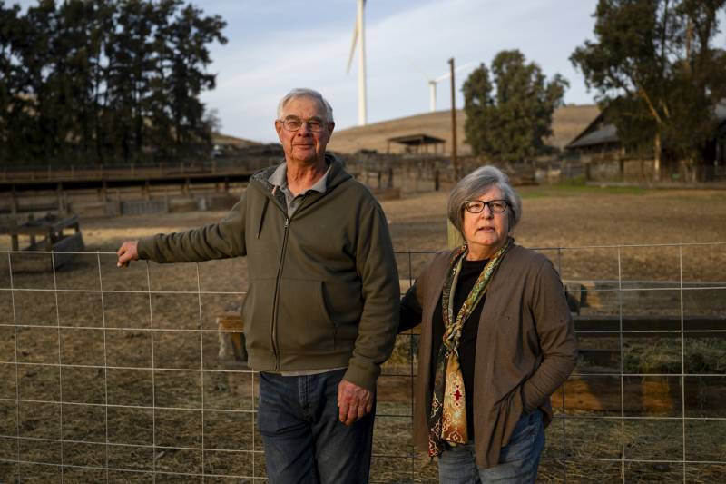 An older white man and an older white woman, stand on farmland looking at the camera.