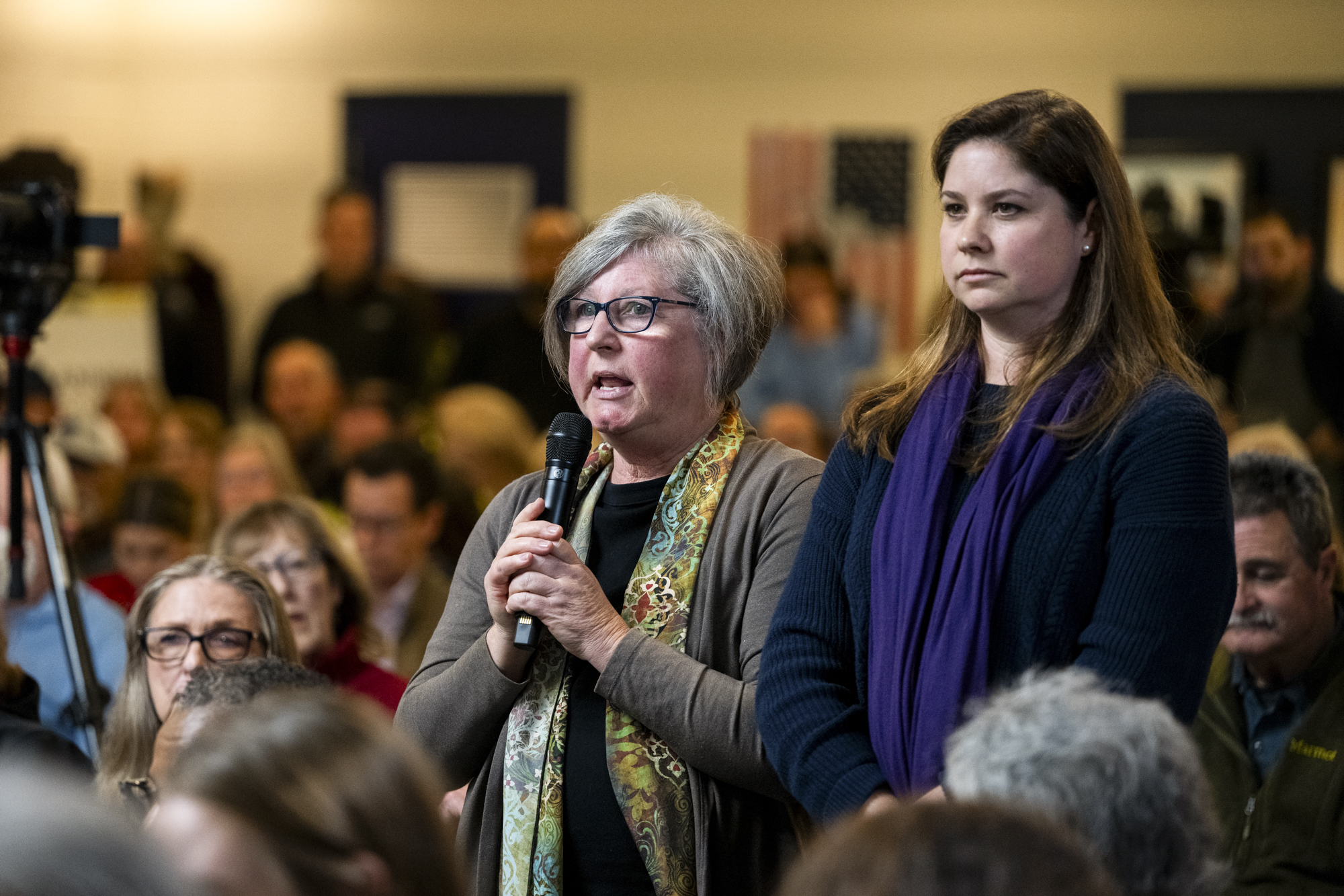 An older white woman and a younger white woman to her left during a town hall, the older woman is speaking into a microphone.