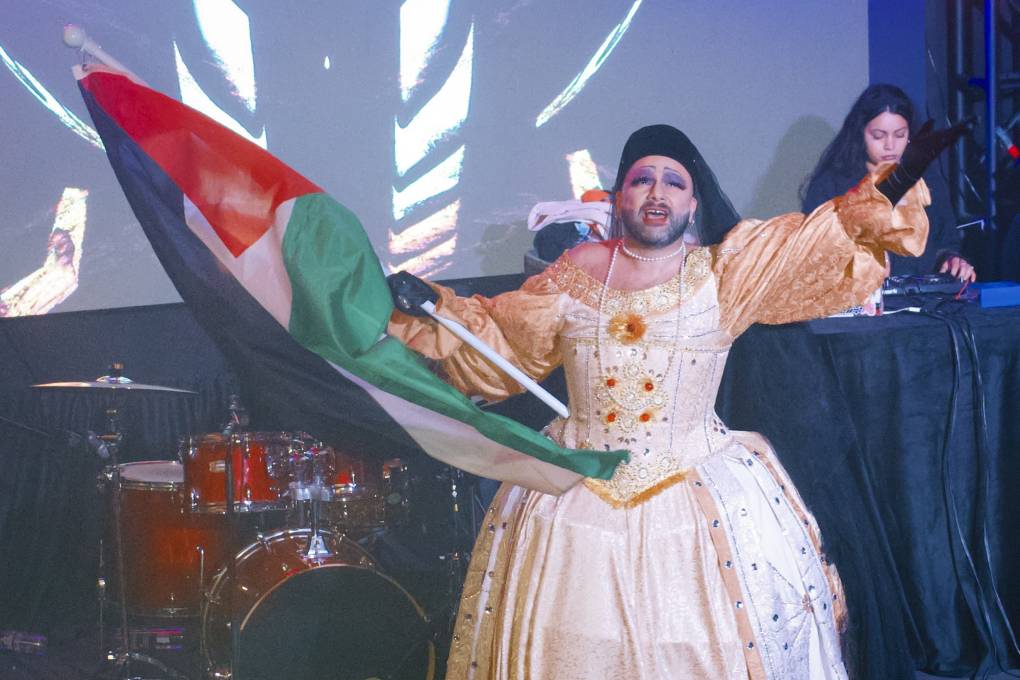 A person with a beard and wearing a dress and a long necklace holds the Palestinian flag on a stage.