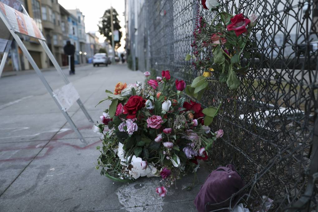 A bouquet of flowers lays on the sidewalk beside a chainlink fence in an urban setting.