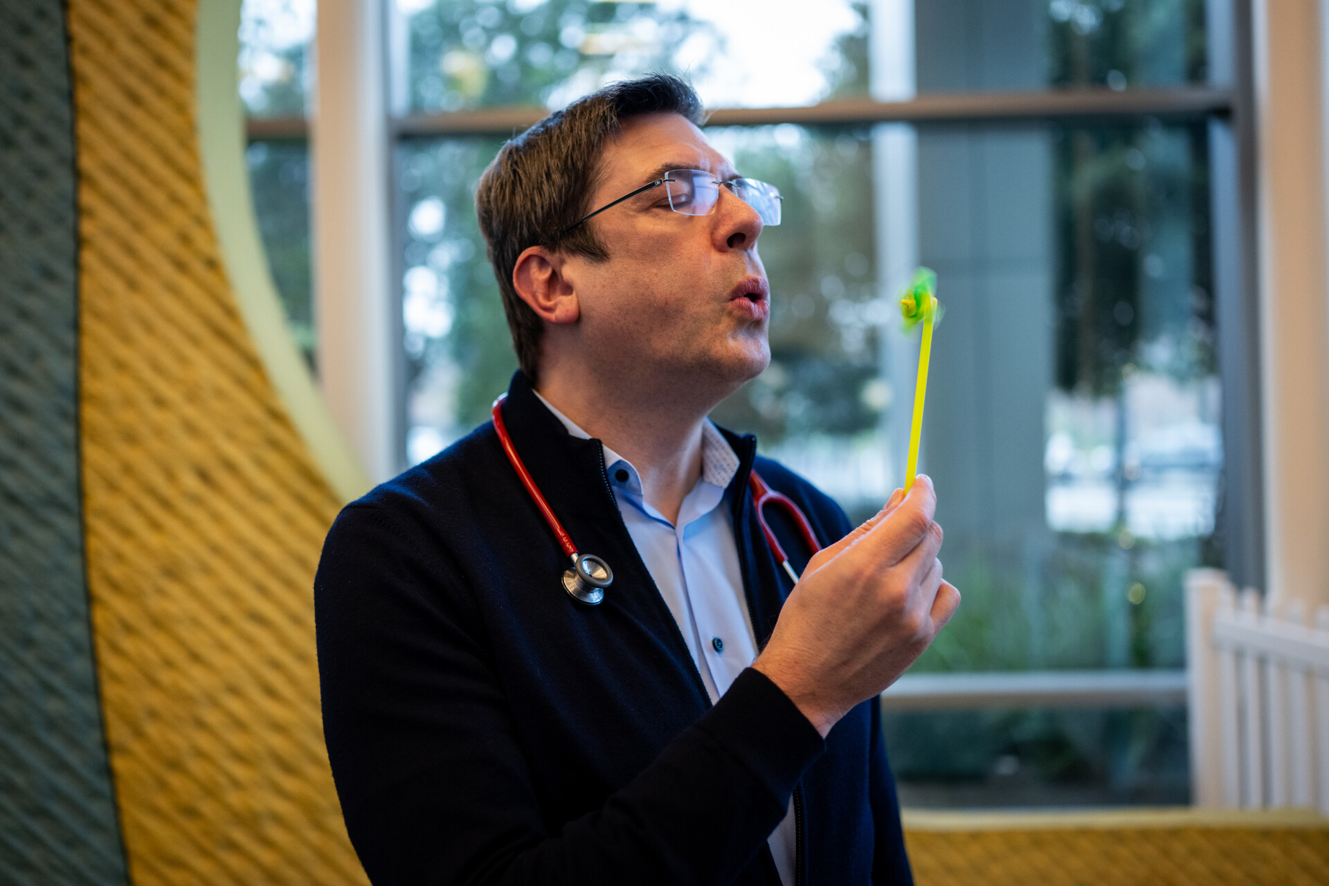 A man with glasses and a stethoscope blows on a mini pinwheel