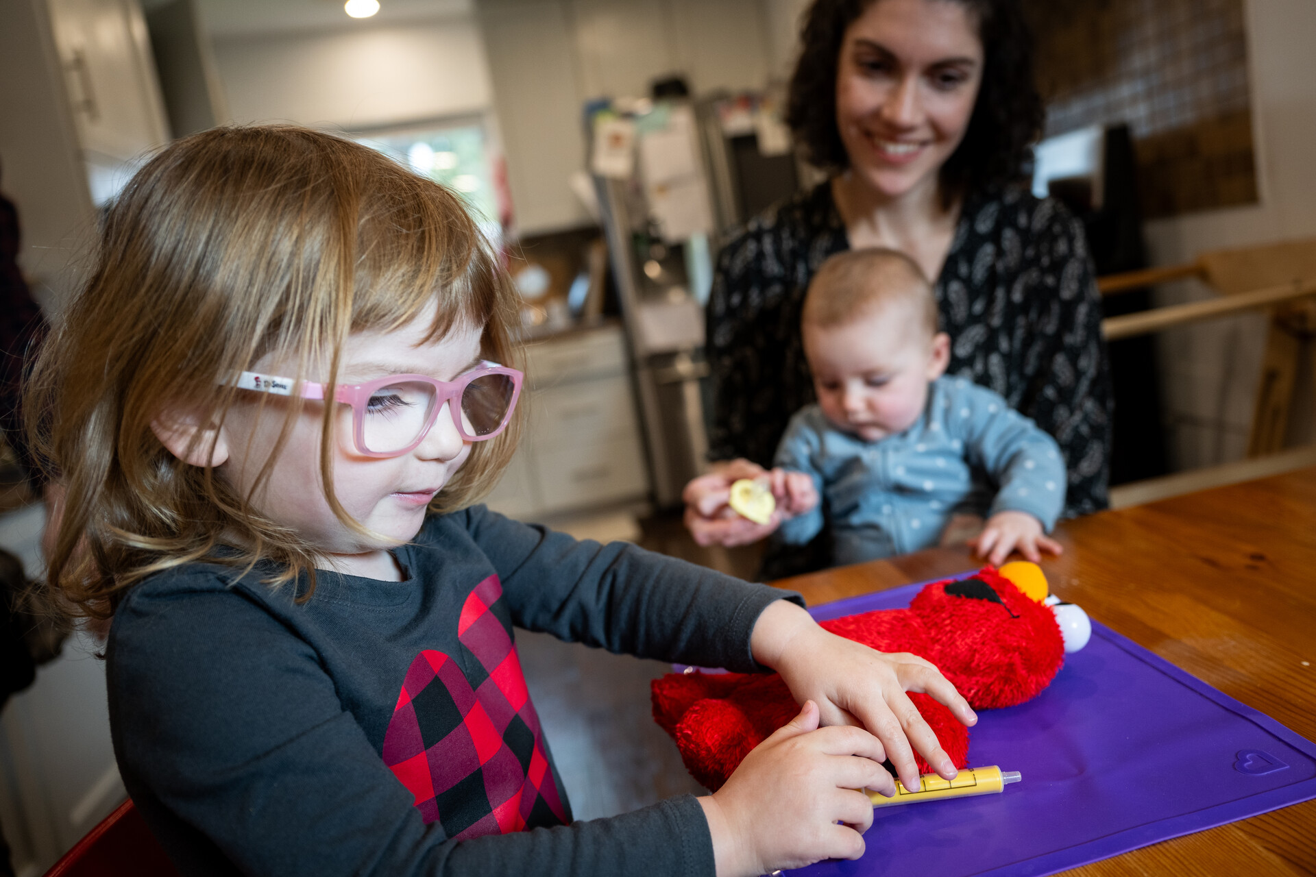 A mother with a baby on her lap sits next to a 3-year-old, who is playing with a toy needle and an Elmo doll.