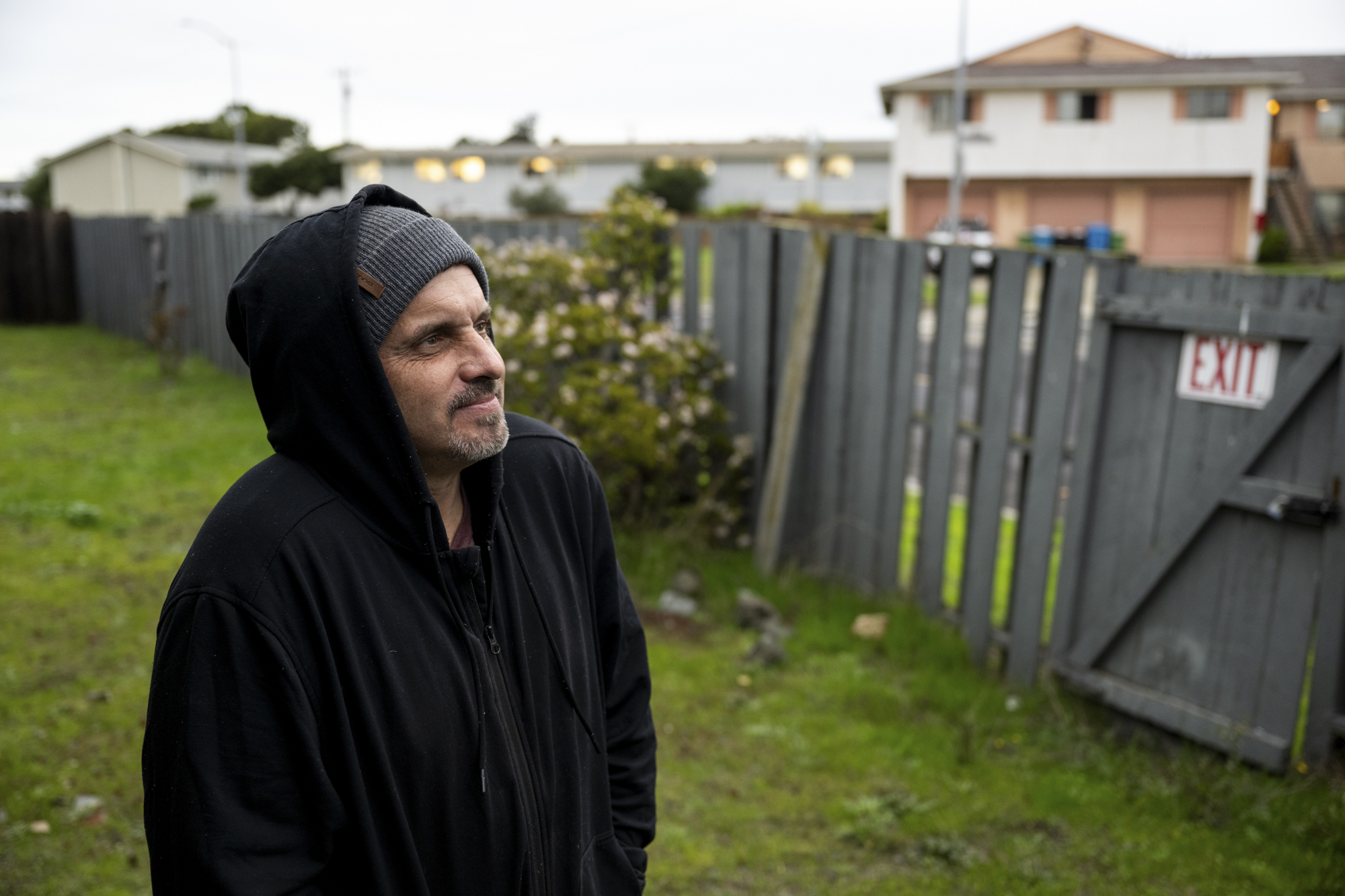 A person wearing a hood stands beside a fence in a residential area.