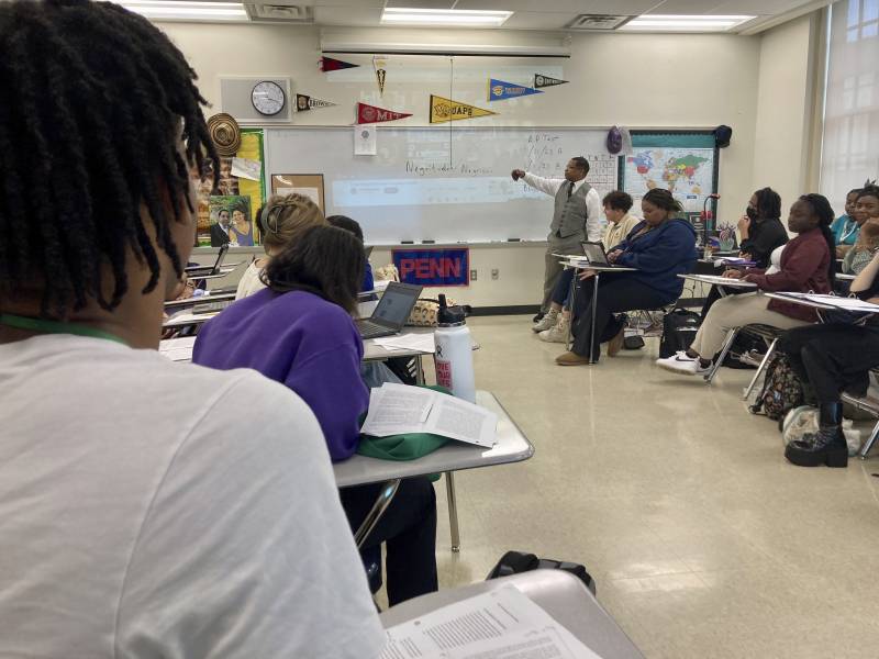 Students sit at desks in a classroom with an adult standing up and pointing at a whiteboard.