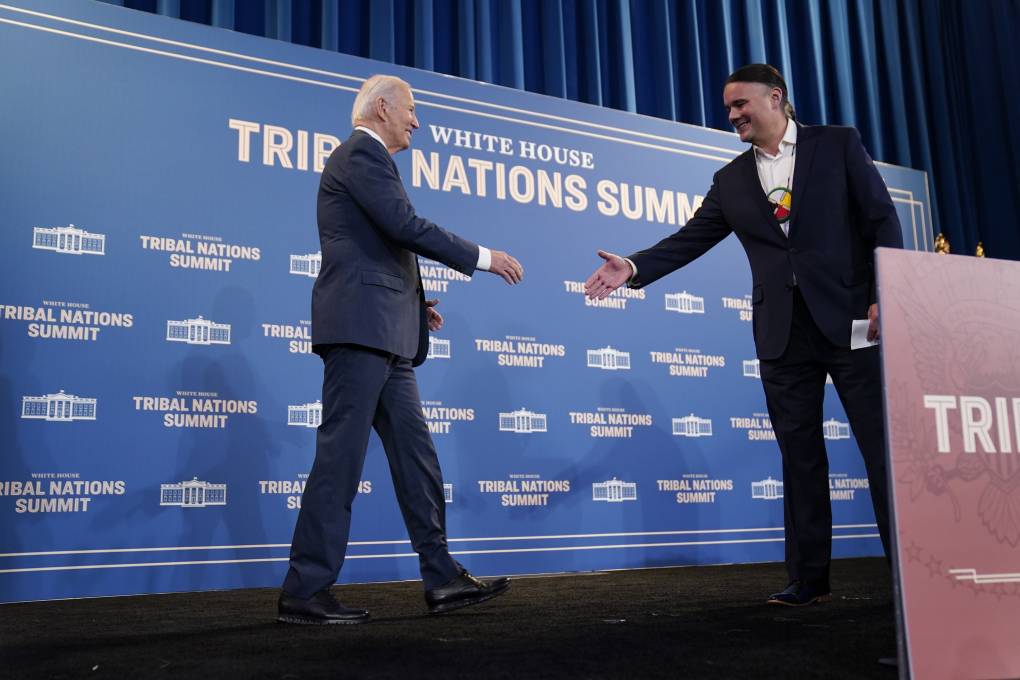 President Biden, on a stage, moves to shake hands with a younger man. Behind them, a sign reads: 'Tribal Nations Summit.'