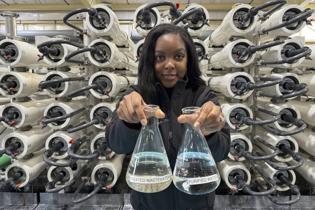 A Black woman holds up two flasks of water behind machinery.