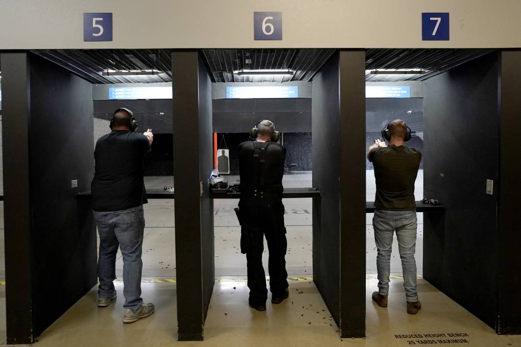 Three men in separate booths wearing headphones face a wall with target practice materials.