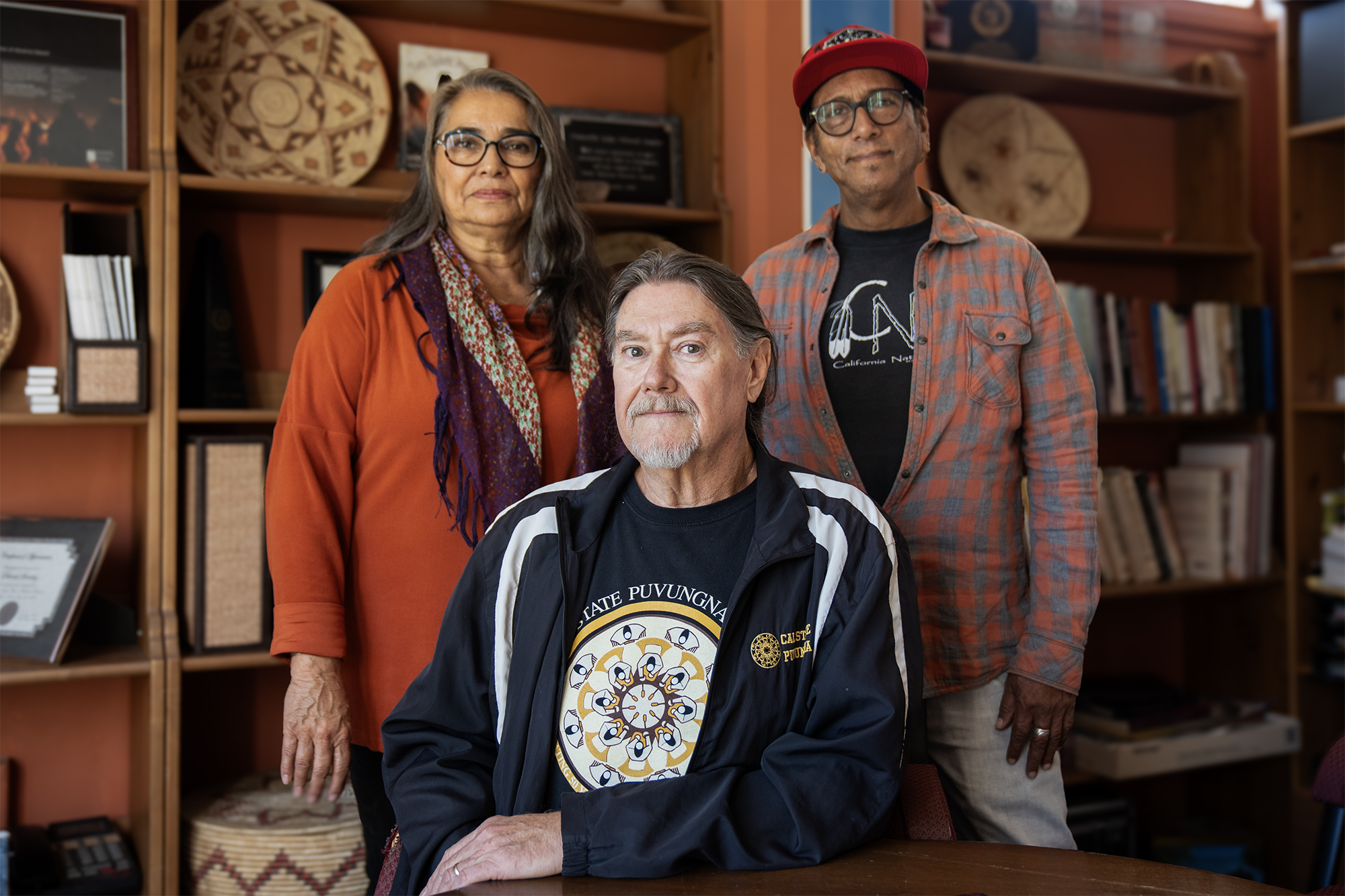 A photo of three individuals are pictured: two men and one woman. All have serious faces as they stand in front of a wall full of bookshelves that have various Native American artifacts and books on it.