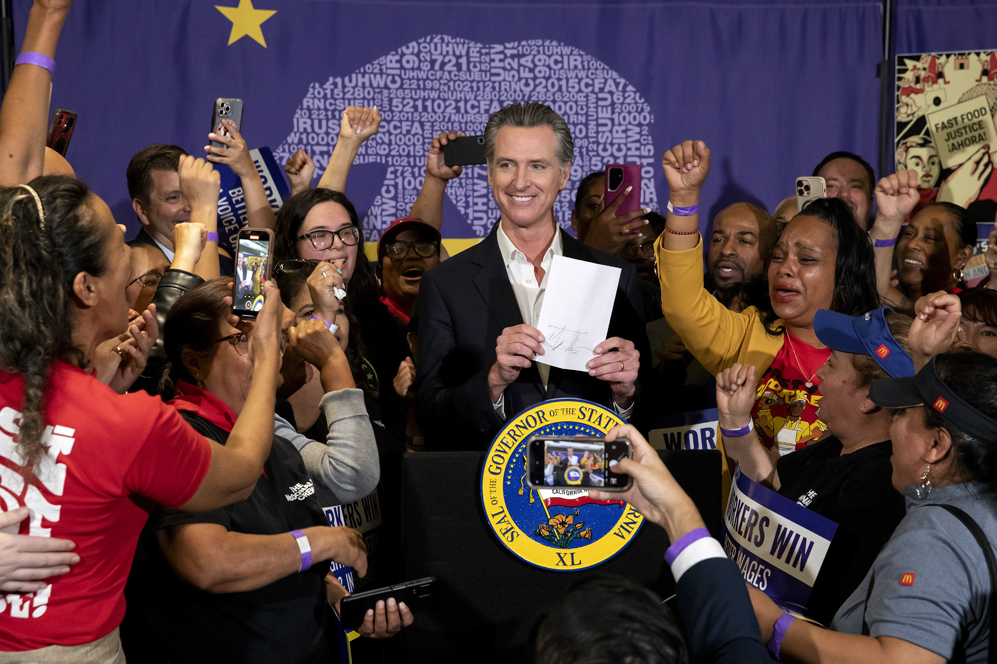Gov. Gavin Newsom is speaking at a podium, smiling, surrounded by press and cheering people.