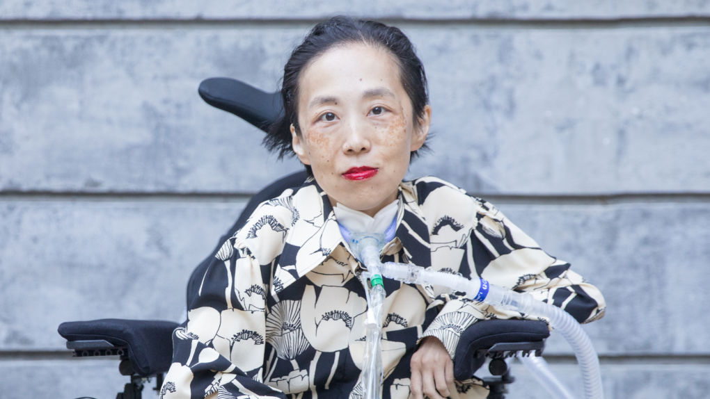 An Asian American disabled woman sits in a wheelchair with a tracheostomy with a tube attached to her ventilator. She is wearing a bold red lip color and a black and beige shirt with a pattern of poppies.