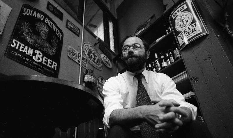A black and white photo of a white bearded man wearing a white button down and a tie. He is seated in front of beer paraphernalia. 
