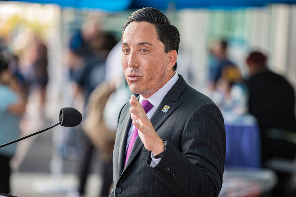 A Latino man in a dark gray suit with a maroon tie gestures as he speaks into a microphone.