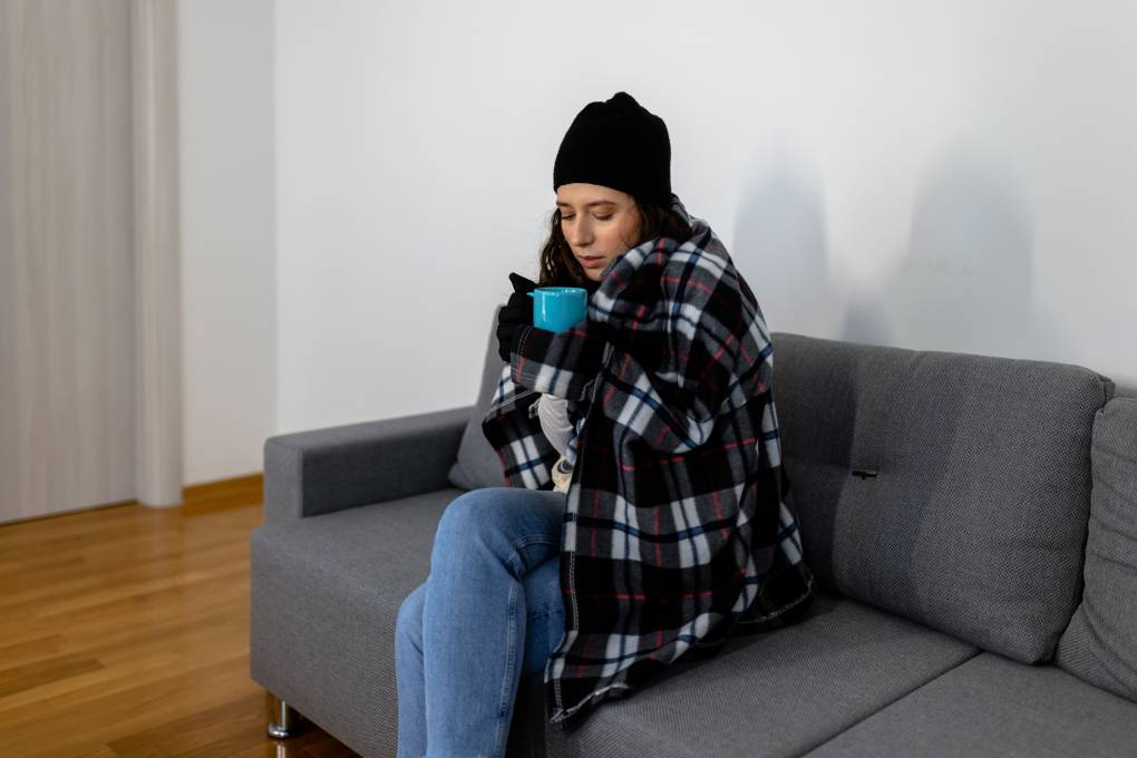 A young woman wearing a winter hat, wrapped in a blanket, sits on a couch, holding a mug.