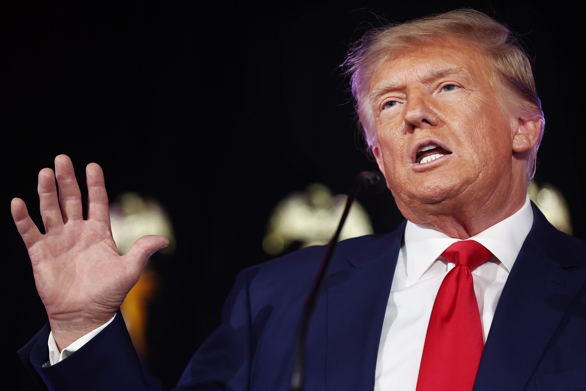An older white man with a navy blue suit and red tie gestures as he speaks.