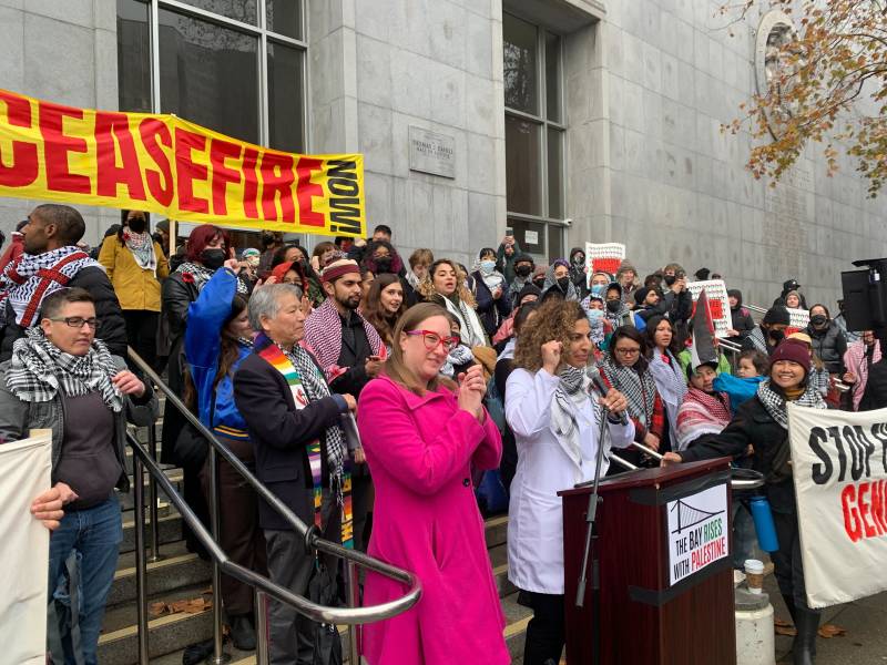A group of people gather for a rally on the steps in front of a courthouse, speaking into a microphone, with a yellow banner that read' Ceasfire Now!' behind them.