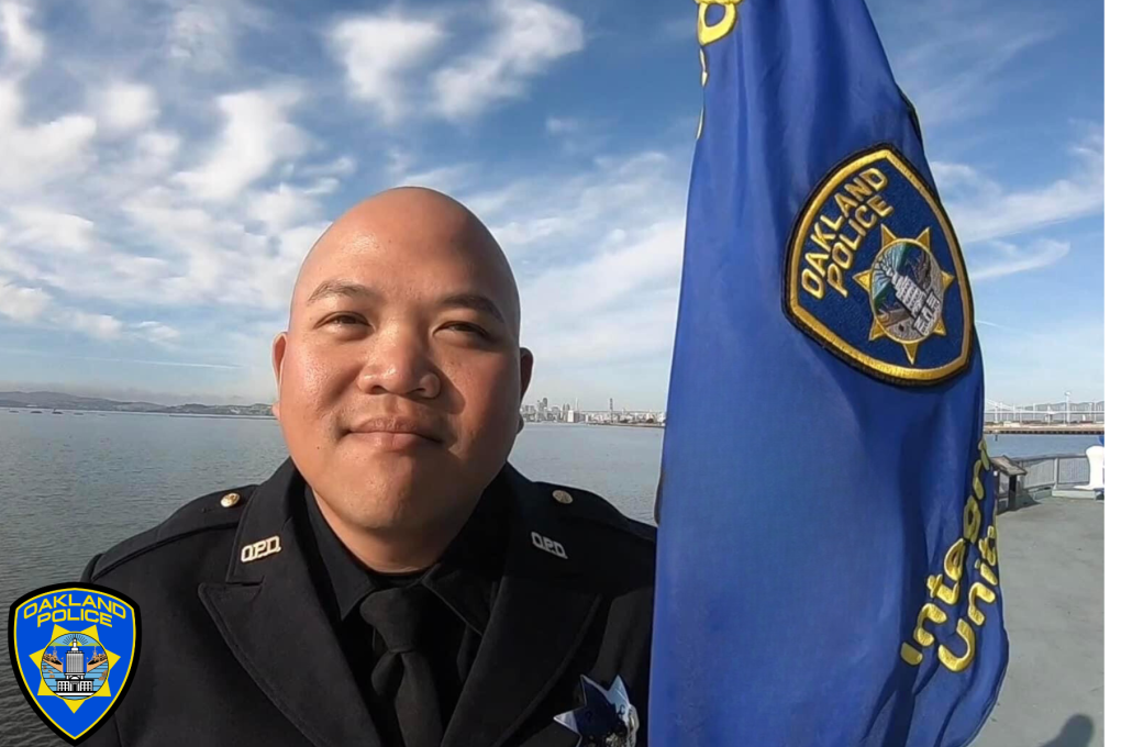 A close-up headshot of a young Asian Oakland police officer next to a blue OPD flag, with a large body of water in the background.