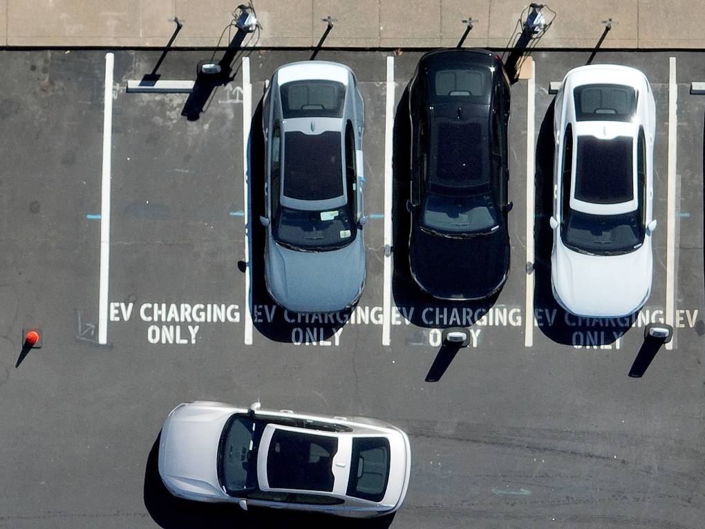 An aerial view of a parking lot that has EV chargers, with three cars parked in 'EV Charging Only' spots, and a fourth car pulling in to another one.