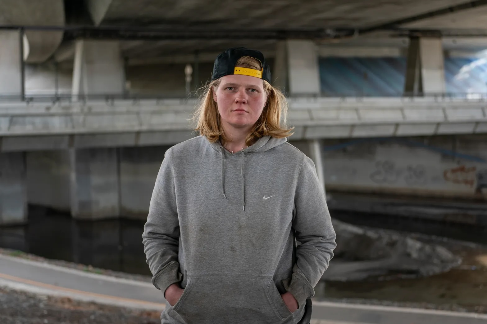 A young white girl with baseball cap worn backward and neck-length blond hair and blue eyes wearing a grey hoodie looks straight in the camera underneath an overpass.
