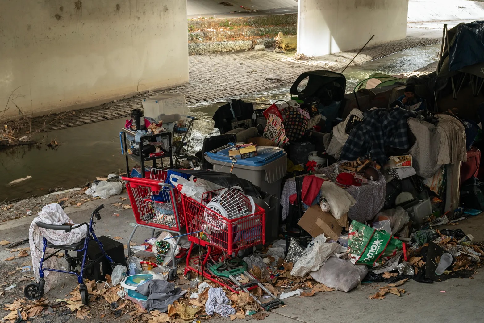 A homeless encampment under a bridge with discarded items strewn around.