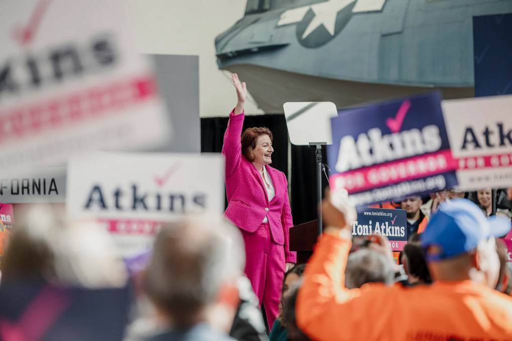 A woman in a pink suit waves and smiles amid a cheering crowd holding banners that read "Atkins."