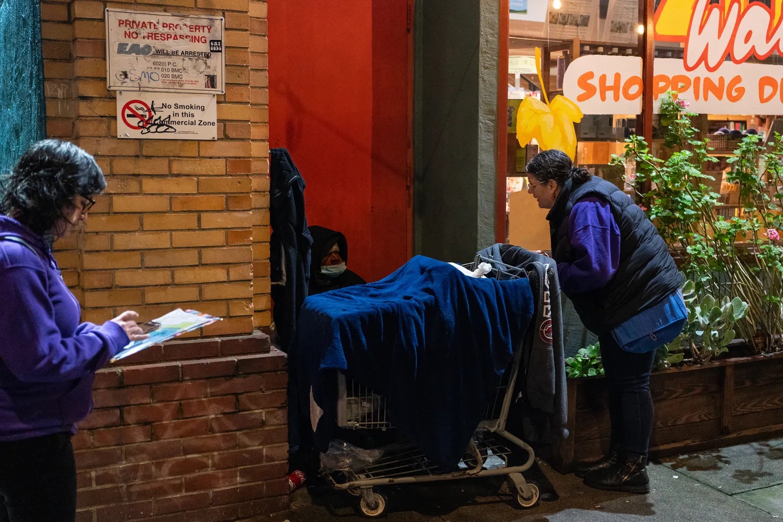 two women talk to a man sitting behind a shopping cart with belongings in it