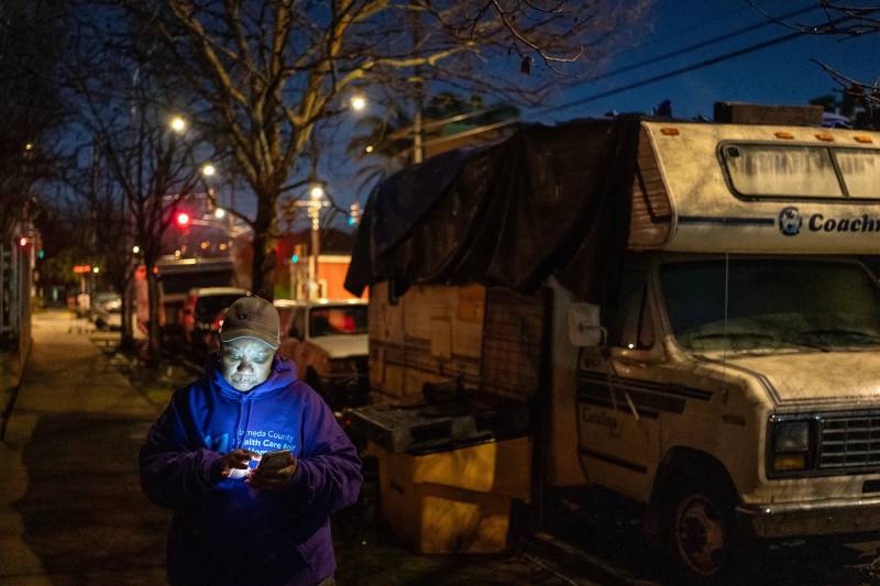 A Black woman in a sweatshirt stands outside an RV on a street, looking at her phone in the dark.