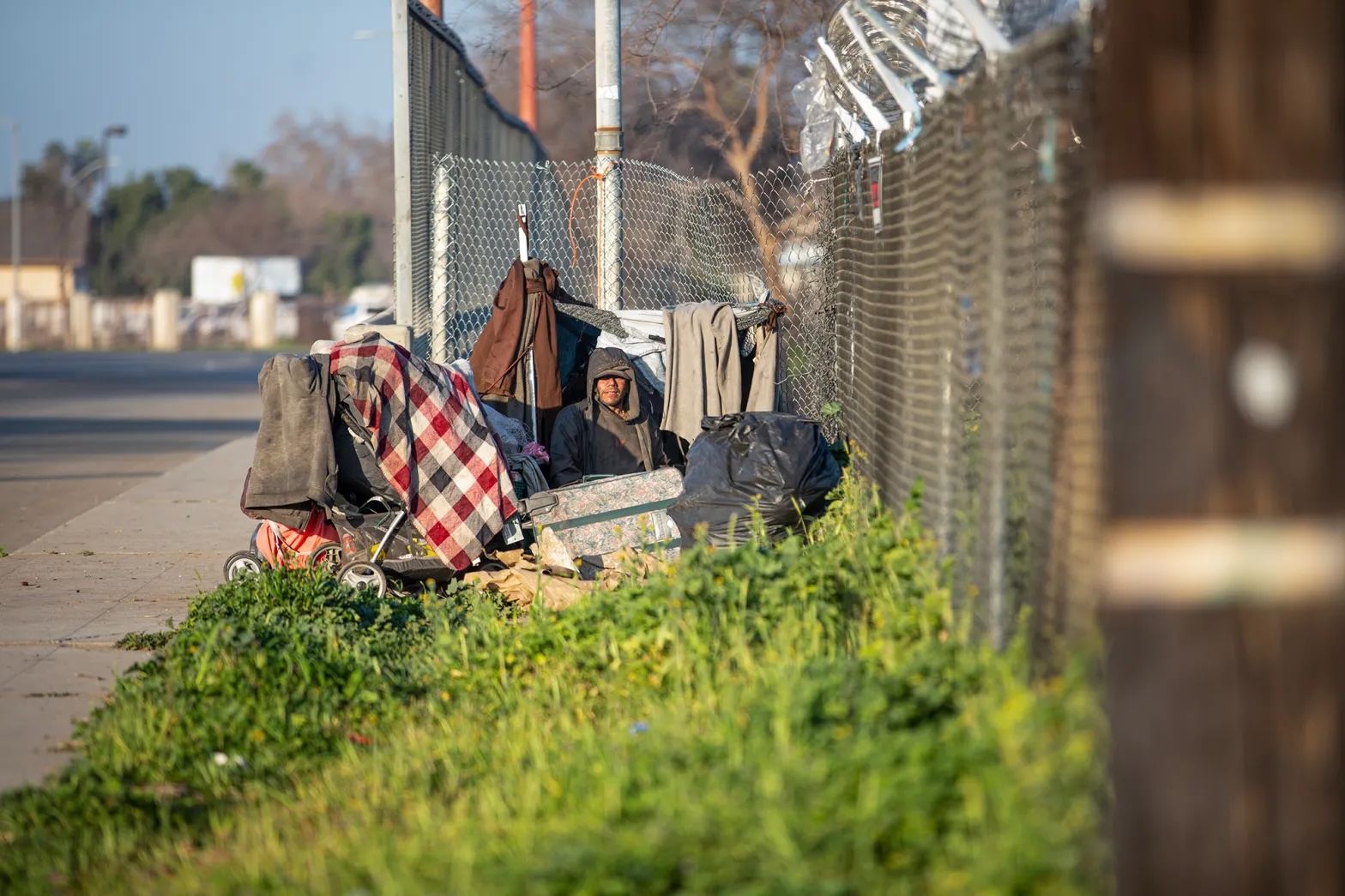 An unhoused person sitting beside a fence with a hoodie pulled over the head and belongings around.