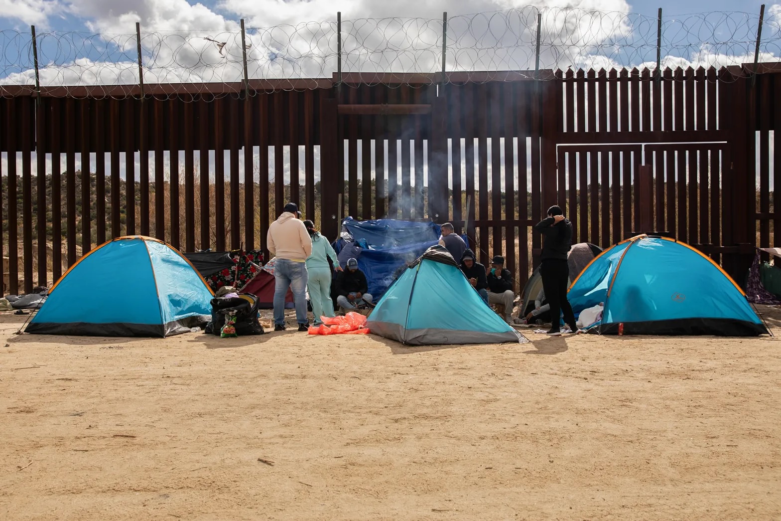 Migrant tents beside a border wall with some people standing and seated between the tents.