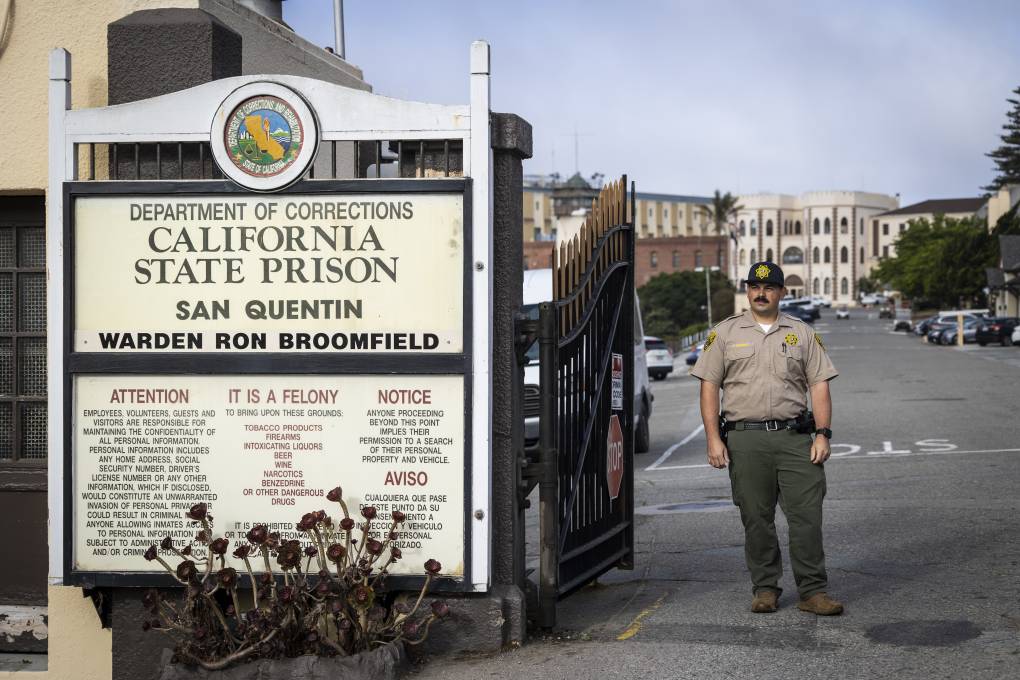 A prison guard in uniform stands in front of a gate with a building in the background.
